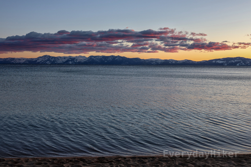 Lake Tahoe at sunset. Pink clouds lead down to an orange hue on the horizon over distant snow capped mountains.  A thin line of the sandy beach may be seen lining the bottom of frame at the edge of the lake.